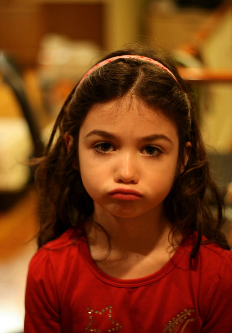 Picture of a little girl in a red shirt with long hair, a pink hair band, and a goofy, annoyed look on her face..
