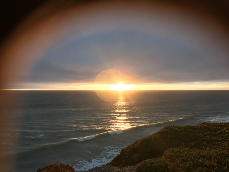 Picture of the ocean with the sun setting and a hazy ring around dit.